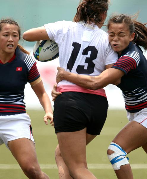 3 rugby players are seen mid match, one player has the rugby ball and a second player is seen mid tackle with the third player standing further in the background